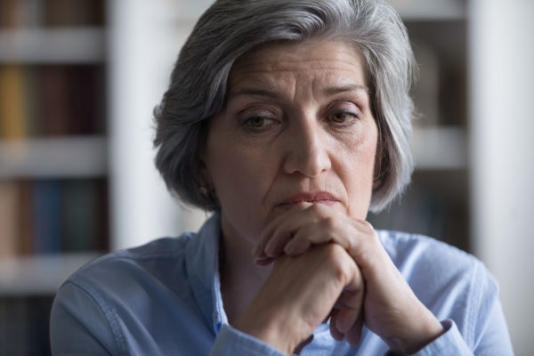 A senior woman with grey hair holds her hands to her mouth as she worriedly looks down and to the side. 