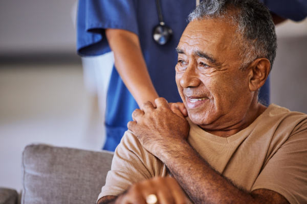 Older man sits on couch as a nurse gently places her hand on his shoulder. 
