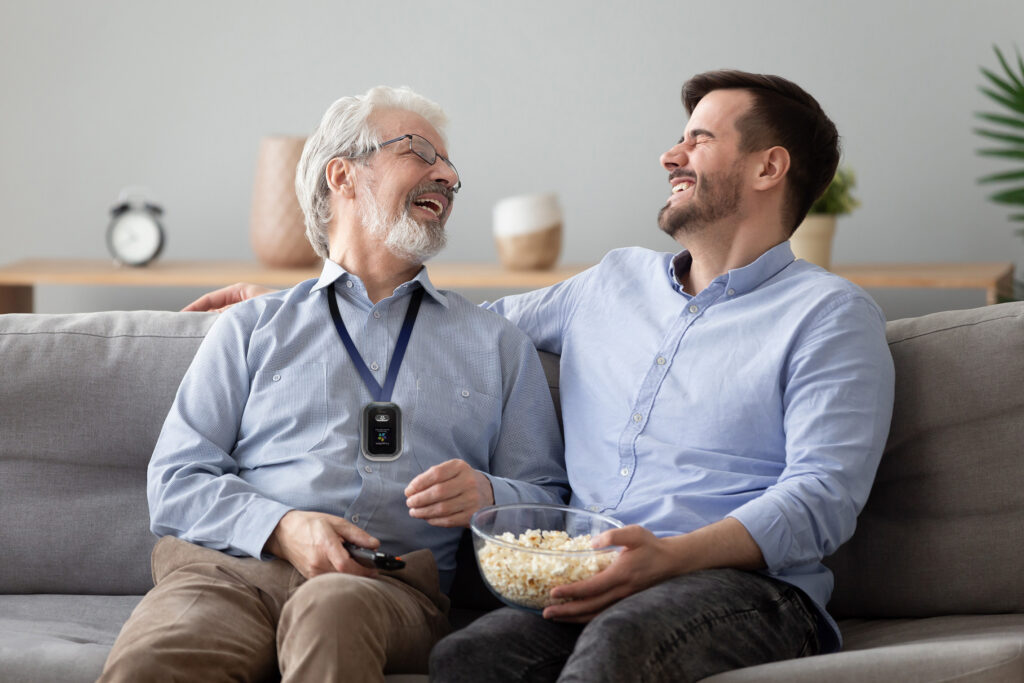 A senior man and young man enjoy popcorn together on the couch as they laugh. 