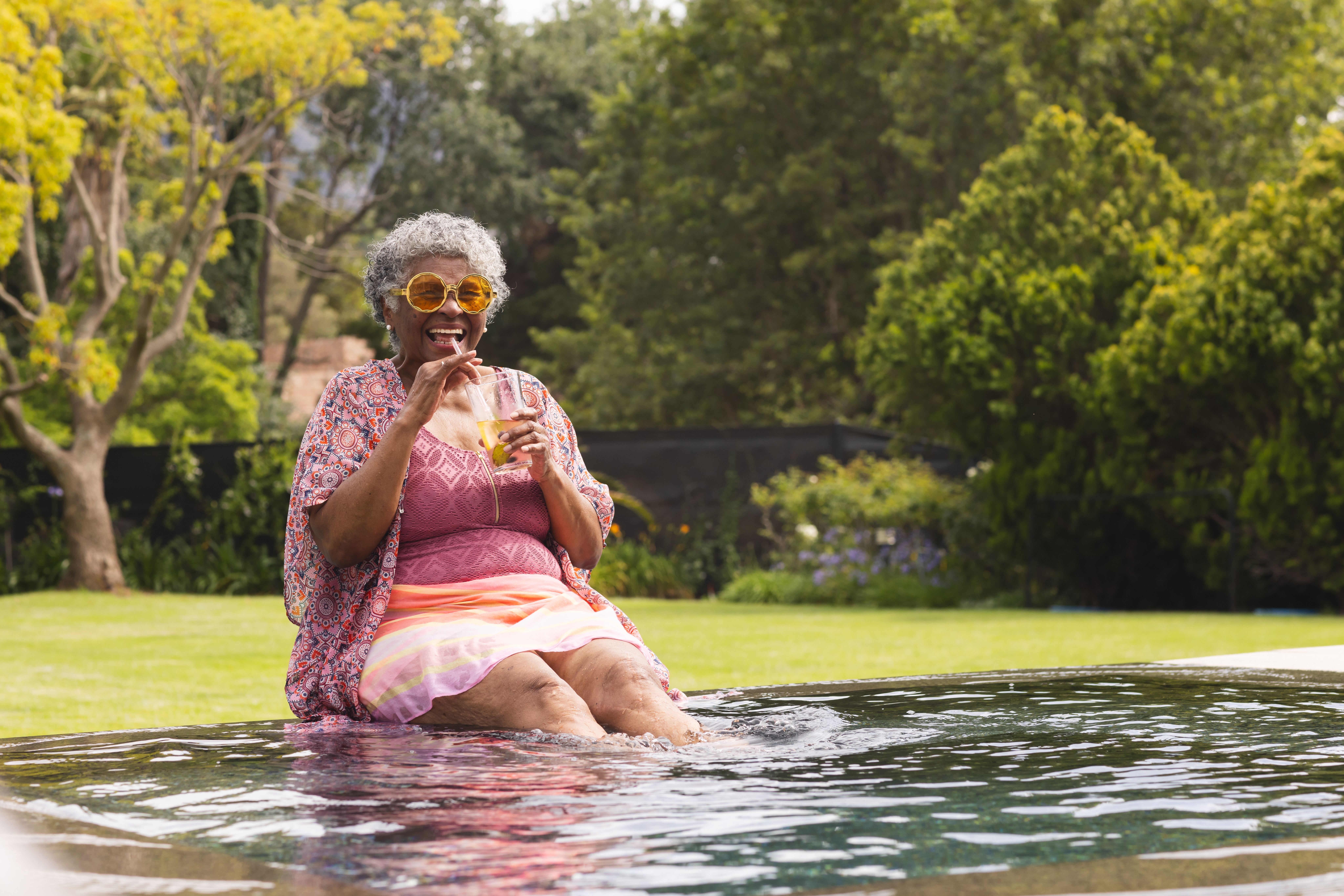 A senior woman smiles while she sips on a drink with her feet in the water.