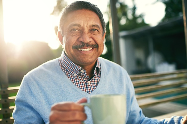 A senior man smiles as he sits on a bench outside while holding a mug.